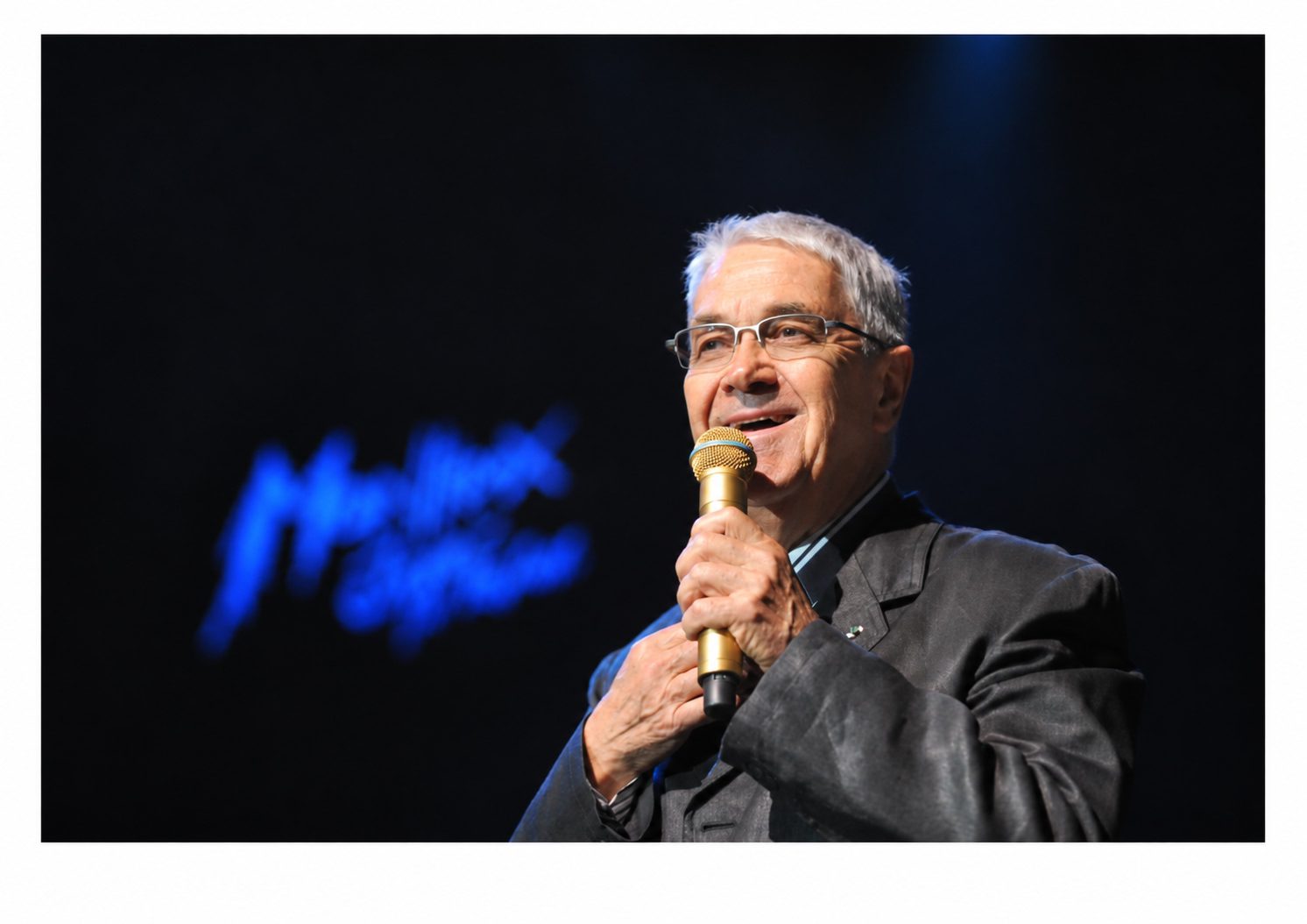 Claude Nobs, Gründer des Montreux Jazz Festival, spricht auf der Bühne in ein goldenes Mikrofon, im Hintergrund leuchtet blau das Logo des Montreux Jazz