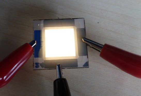 Warmweiss leuchtendes OLED-Panel des OSRAM-Demonstrators, mit zwei roten Krokodilklemmen kontaktiert